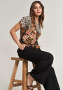 A woman with shoulder-length brown hair and a relaxed expression poses sideways on a wooden stool, wearing a Princess Goes Hollywood Seidenbluse mit floralem Leoprint, schwarze weite Hosen und schwarze Sandalen mit Absatz vor neutralem Hintergrund.