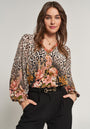 A woman with wavy brown hair wears a Princess Goes Hollywood Seide blouse with V-Ausschnitt and floraler Leoprint in pink, paired with high-waisted black trousers. She stands against a plain light background, smiling slightly.