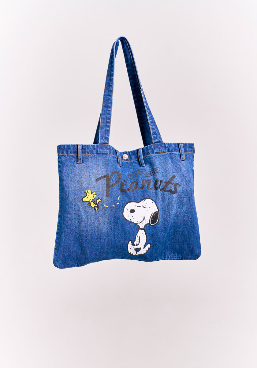A blue denim-shopper tote bag by Princess Goes Hollywood features Snoopy and Woodstock with Peanuts in a playful font above them, celebrating the Peanuts 75th Anniversary. The bag has long handles and a light background behind the characters.