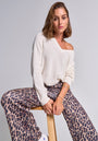 A woman with shoulder-length brown hair sits on a wooden stool against a plain backdrop, wearing a white Princess Goes Hollywood Strickpullover with Wolleanteil and high-waisted, wide-leg leopard print pants, plus bracelets and white Converse sneakers.