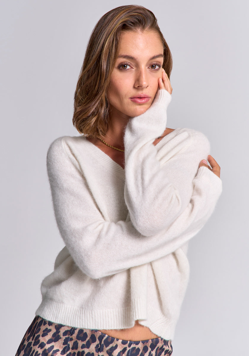 A woman with shoulder-length light brown hair wears a loose, white Strickpullover mit Wolleanteil by Princess Goes Hollywood, long sleeves, a gold necklace, and leopard-print pants, gently touching her face against a light grey background.