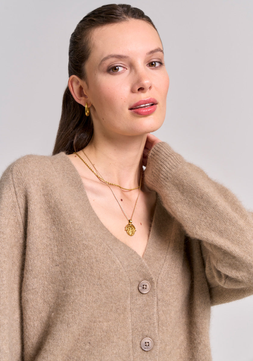 A woman with light skin and brown hair tied back wears a tan Princess Goes Hollywood V-neck Strickjacke with large buttons and gold jewelry—hoop earrings and layered necklaces. She looks at the camera neutrally, her hand near her neck.