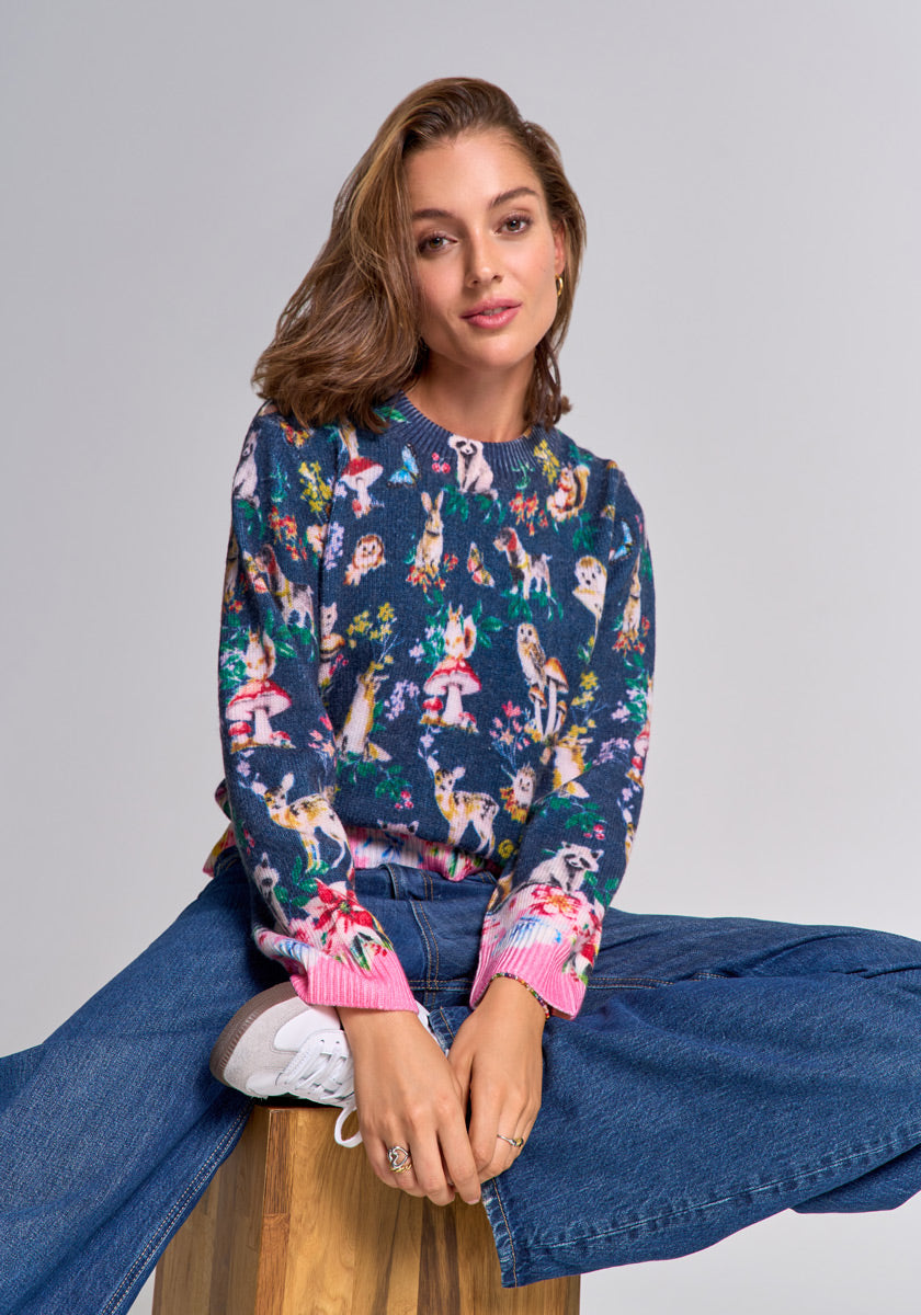 A young woman with shoulder-length brown hair sits on a wooden stool against a gray backdrop, wearing a Princess Goes Hollywood Merino wool sweater with woodland animal print, wide-leg blue jeans, and gray sneakers. She smiles softly at the camera.