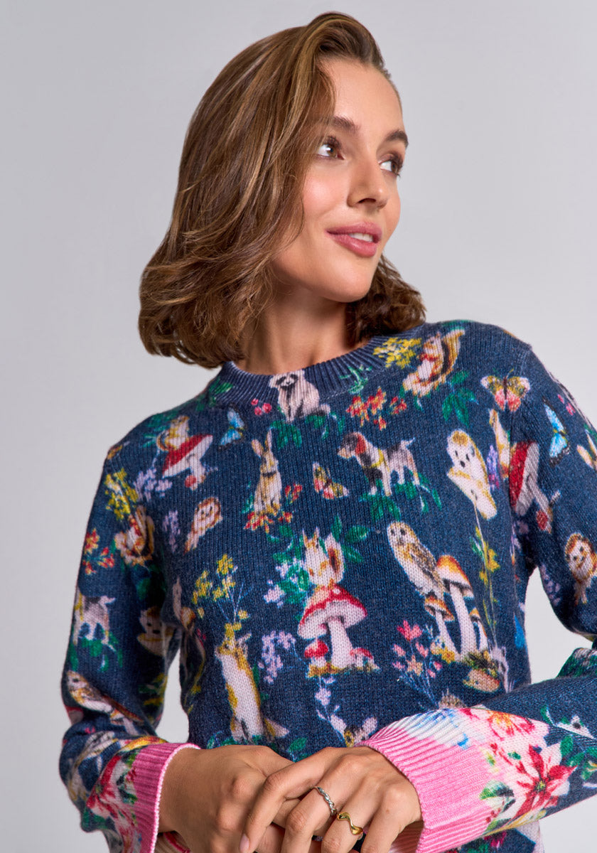 A woman with short, wavy brown hair smiles slightly and looks left. She wears a blue Princess Goes Hollywood Strickpullover with colorful Waldtier-Print, nature illustrations, and bright pink ribbed floral cuffs. Background is plain light gray.