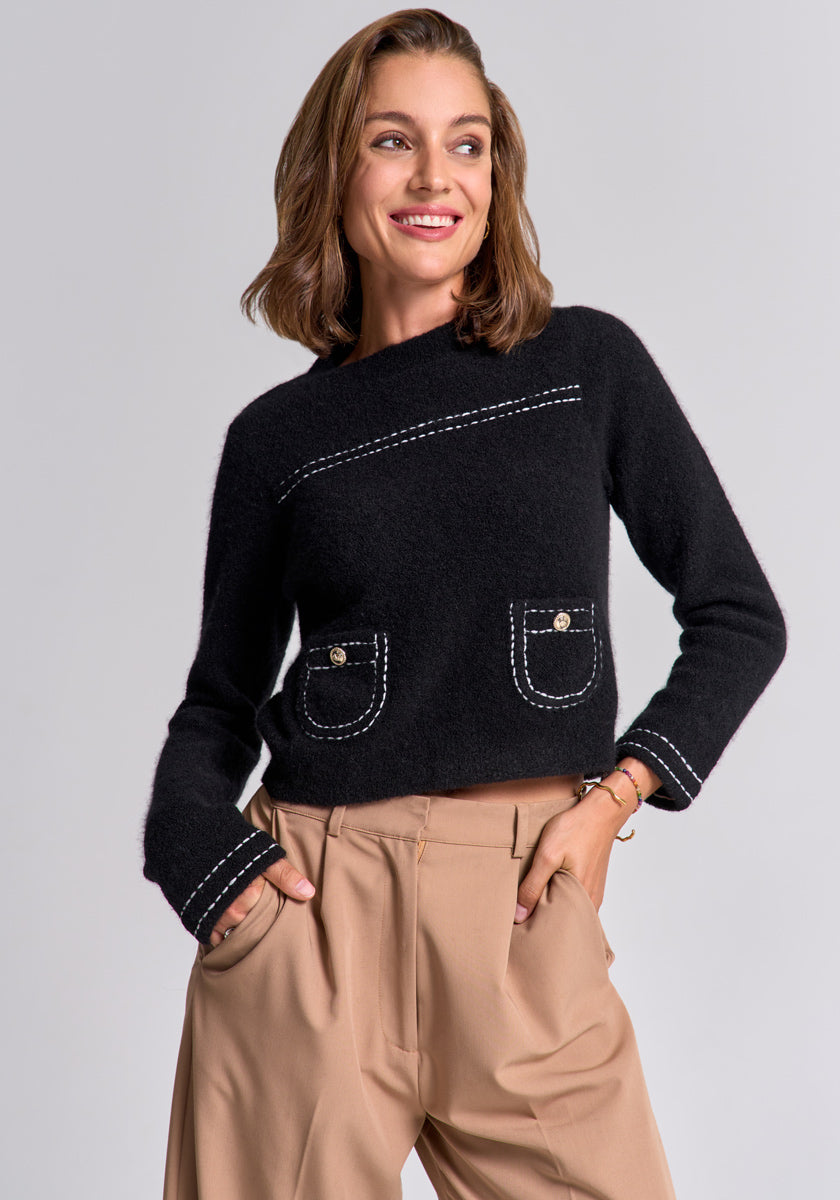 A woman with light brown, shoulder-length hair smiles to the side. She wears a black Princess Goes Hollywood Merino-Kaschmir-Pullover with Rundhalsausschnitt and white stitching, paired with beige high-waisted pleated pants against a plain light gray background.