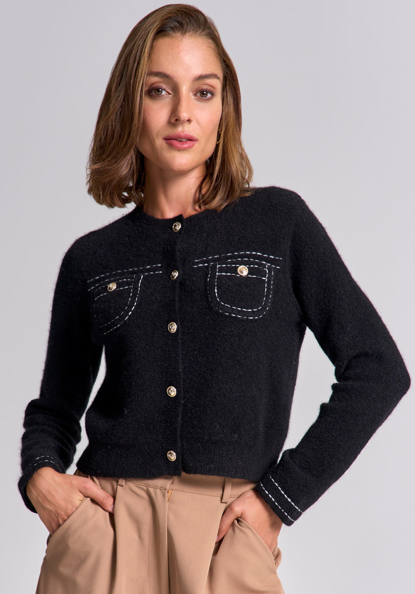 A woman with shoulder-length brown hair wears a schwarze Merino-Kaschmir-Strickjacke mit goldfarbenen Knöpfen und weißer Naht sowie zwei Brusttaschen von Princess Goes Hollywood und hochgeschnittene beige Hosen. Sie steht frontal vor grauem Hintergrund.