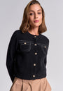 Eine Frau mit schulterlangem, hellbraunem Haar trägt von Princess Goes Hollywood eine schwarze Merino-Kaschmir-Strickjacke mit goldfarbenen Knöpfen und Rundhalsausschnitt sowie beigefarbene, plissierte High-Waist-Hosen.