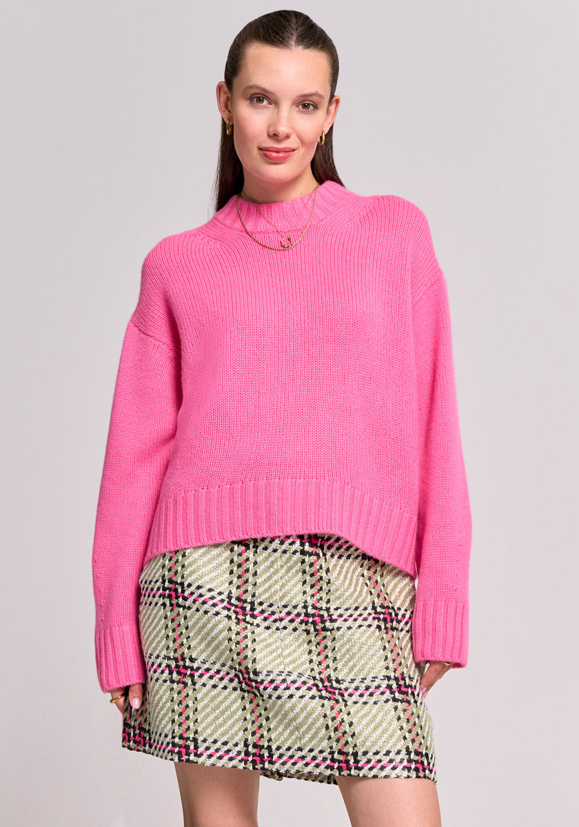 A woman stands against a light grey background, wearing a bright pink Princess Goes Hollywood Kaschmir Pullover mit Stehkragen and a high-waisted green-and-pink plaid skirt. She has straight brown hair pulled back, gold earrings, and smiles subtly with hands at her sides.