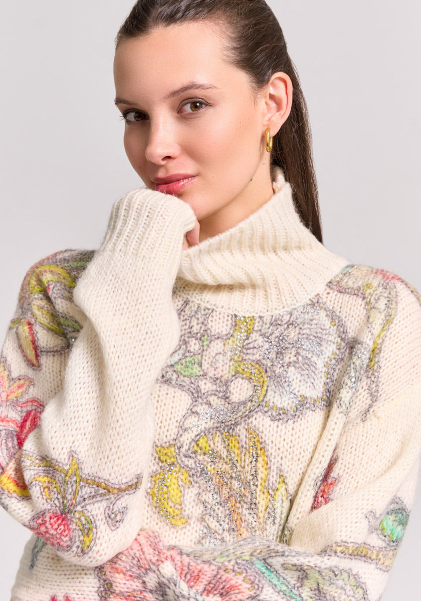 A woman with fair skin and brown hair wears a chunky Princess Goes Hollywood Pullover Wolle-Alpaka-Mix with colorful floral patterns and a cozy roll-neck. She gazes at the camera, chin on hand, small hoop earrings visible against a plain background.