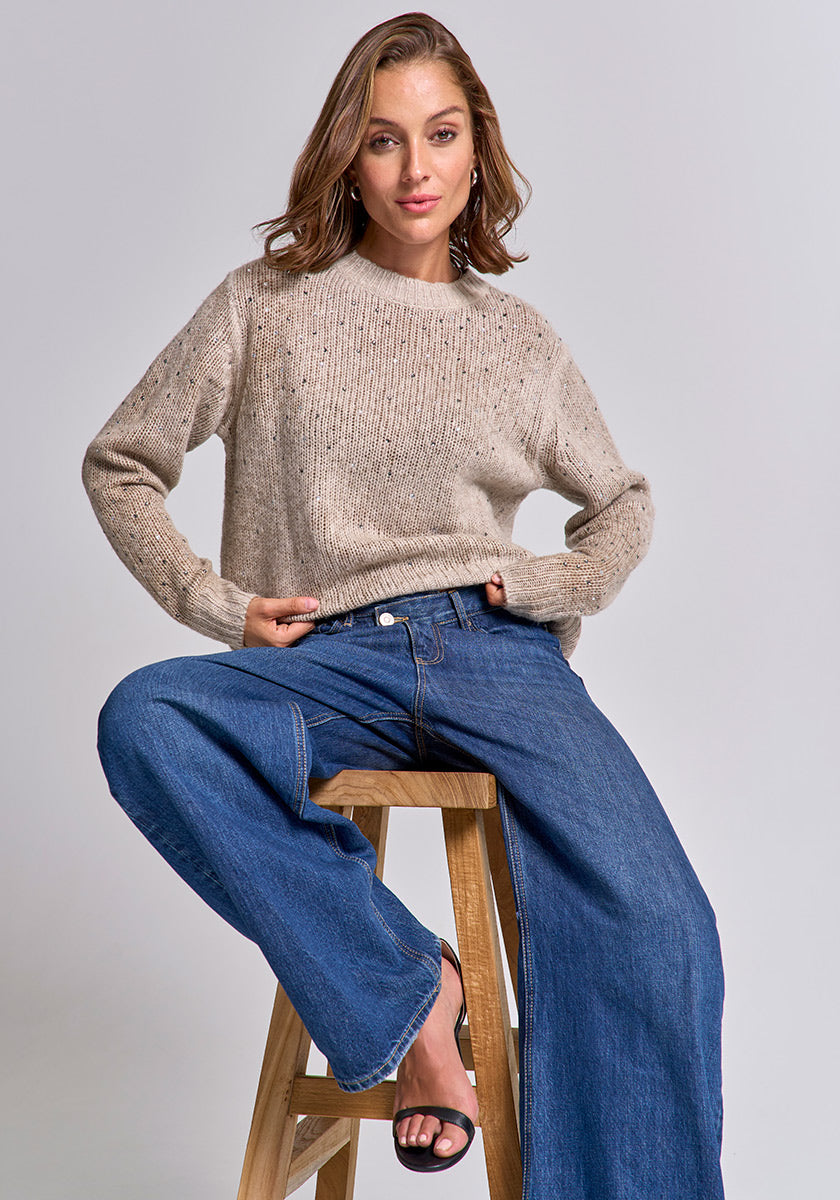 A woman with wavy light brown hair sits on a wooden stool against a plain background, wearing a beige Princess Goes Hollywood Strickpullover Damen, wide-leg blue jeans, and black heeled sandals. She looks at the camera with a slight smile.