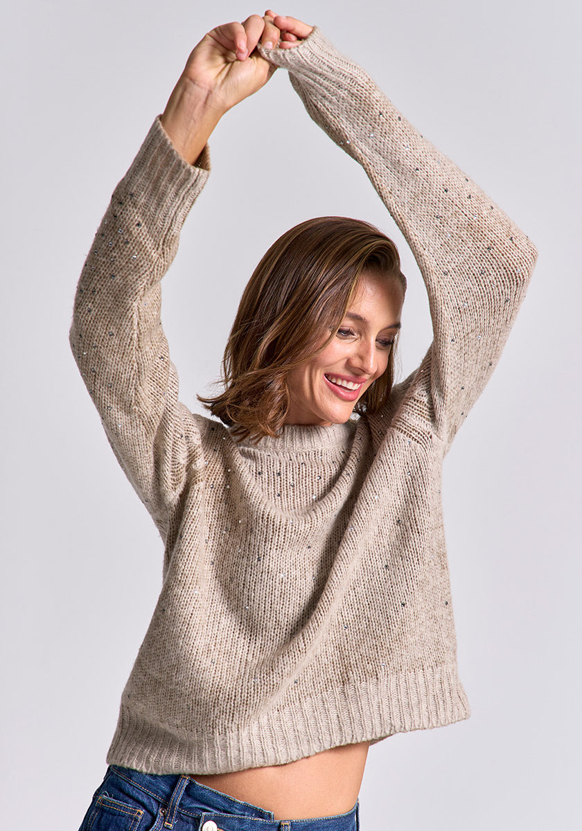 A woman in a loose beige Princess Goes Hollywood Strickpullover Damen and blue jeans raises her arms overhead, smiling with her eyes closed. Her chin-length wavy brown hair and a plain light gray background enhance the relaxed, cheerful vibe.
