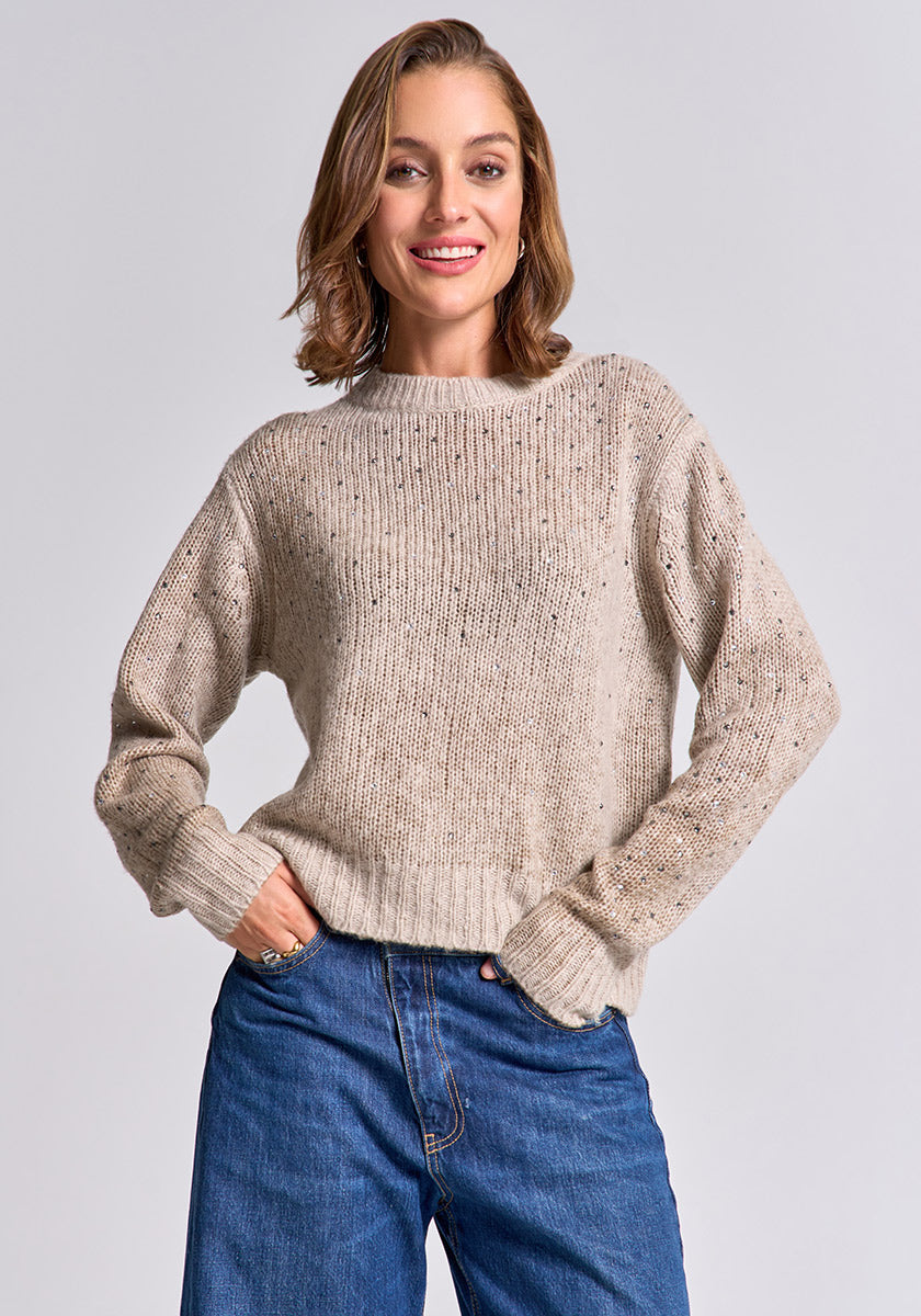A woman with shoulder-length brown hair smiles at the camera, wearing a beige Strickpullover Damen from Princess Goes Hollywood and blue jeans. She stands with one hand in her pocket against a plain light gray background.