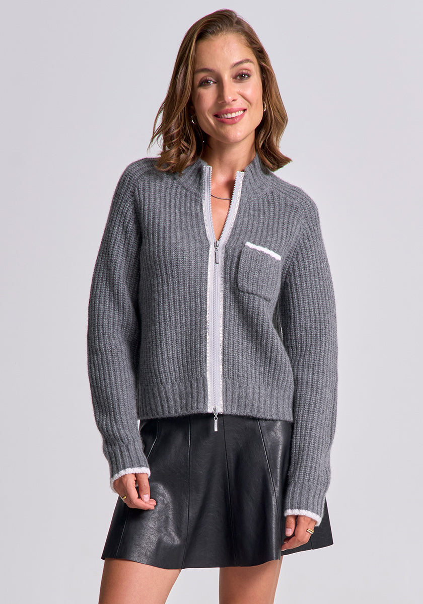 A woman with short brown hair smiles, wearing a Princess Goes Hollywood gray Wolle-Kaschmir-Mix Strickjacke with a white-trimmed chest pocket and black leather mini skirt, standing against a plain light gray background.