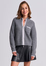 A woman with short brown hair smiles, wearing a Princess Goes Hollywood gray Wolle-Kaschmir-Mix Strickjacke with a white-trimmed chest pocket and black leather mini skirt, standing against a plain light gray background.