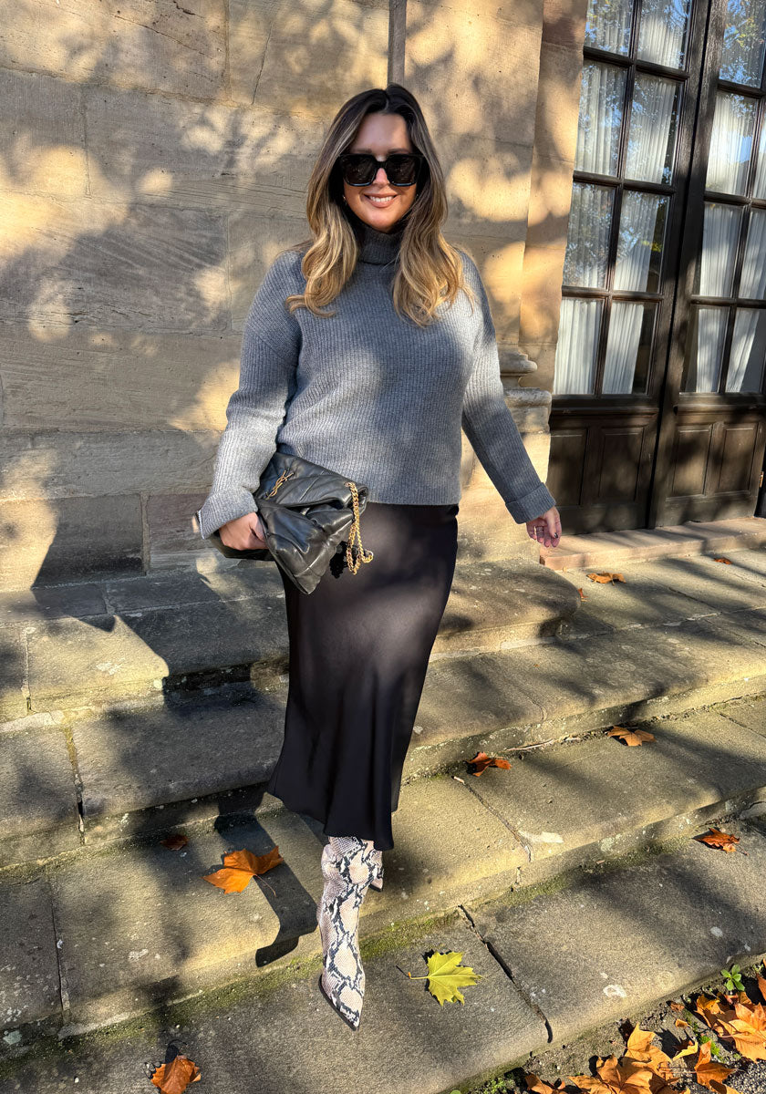 Eine lächelnde Frau in einem grauen Princess Goes Hollywood Rollkragen-Strickpullover, einem schwarzen Midirock, Schlangenlederstiefeln und einer Sonnenbrille steht auf einer Steintreppe mit Herbstlaub. In der Hand hält sie eine schwarze, gesteppte Handtasche mit Kettenriemen.