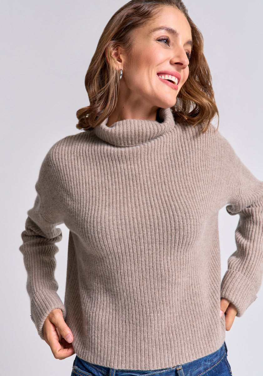 A woman with light brown hair wears a beige Princess Goes Hollywood wool-cashmere blend turtleneck sweater and blue jeans, smiling as she looks right with one hand on her hip, against a plain light gray background.