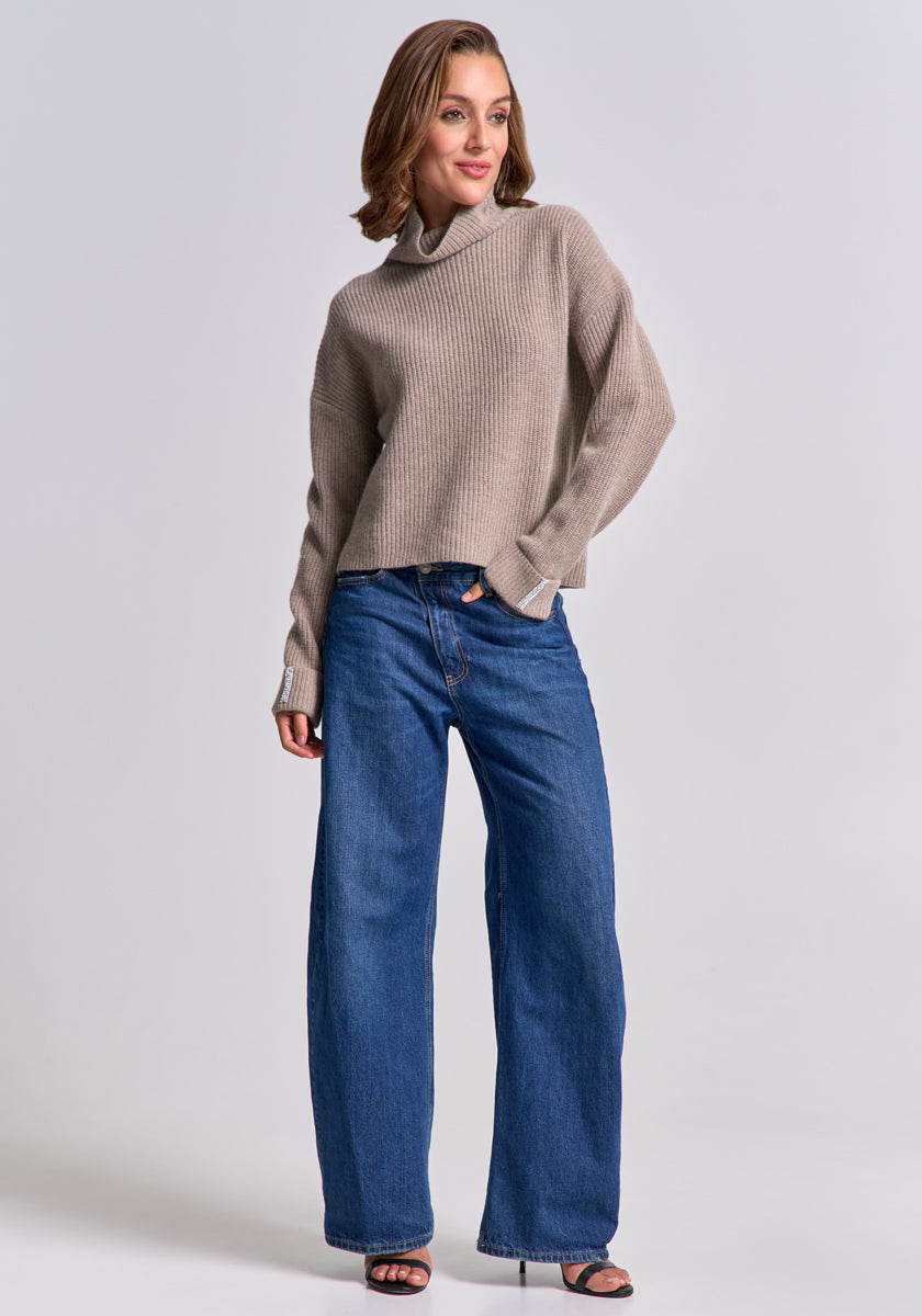 A woman with light brown wavy hair smiles against a plain light gray background, wearing a beige Princess Goes Hollywood Wolle-Kaschmir-Mix Strickpullover, relaxed blue wide-leg jeans, and black open-toe sandals.