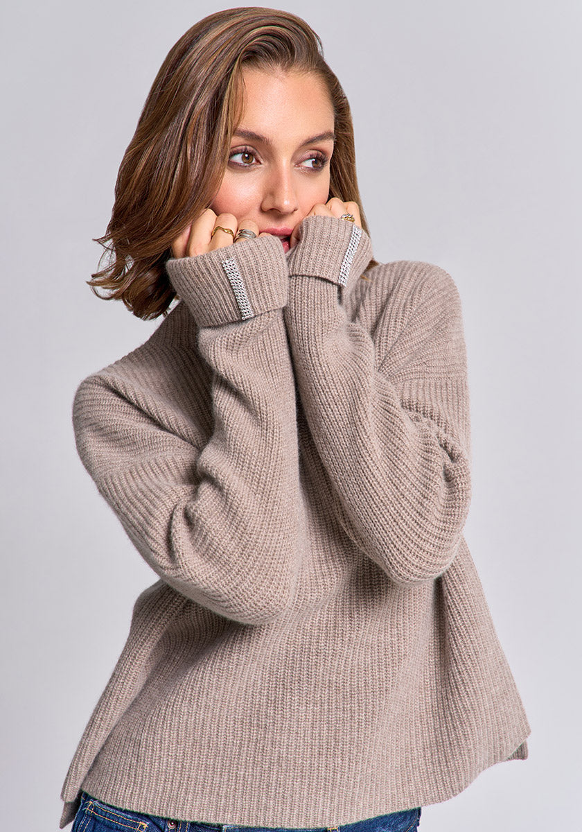 A woman with light brown hair wears a loose, beige Princess Goes Hollywood wool-cashmere blend sweater with long sleeves covering her hands. She gazes thoughtfully to the side, gently holding the collar near her face against a plain light gray background.