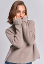 A woman with light brown hair wears a loose, beige Princess Goes Hollywood wool-cashmere blend sweater with long sleeves covering her hands. She gazes thoughtfully to the side, gently holding the collar near her face against a plain light gray background.