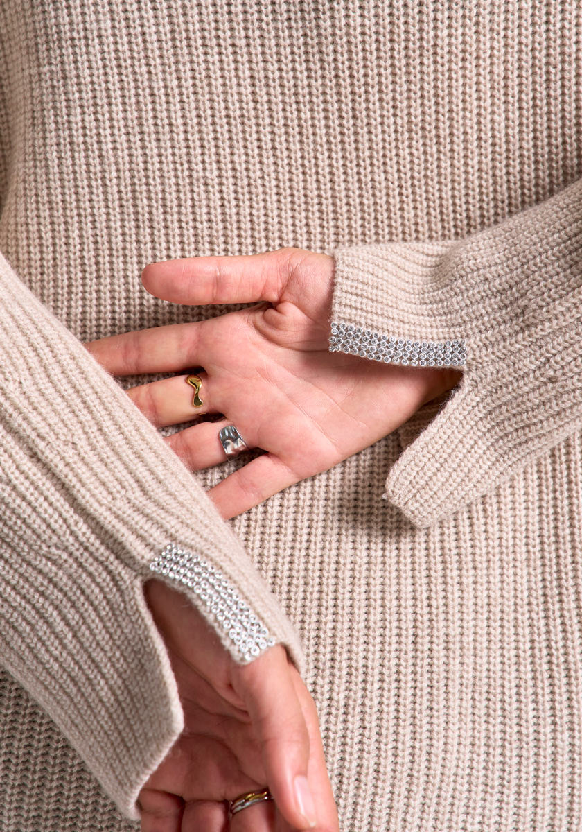 Close-up of two hands against a beige Princess Goes Hollywood wool-cashmere sweater with small cuff slits and silver detail. The person wears a gold ring and a wavy silver ring on their fingers.