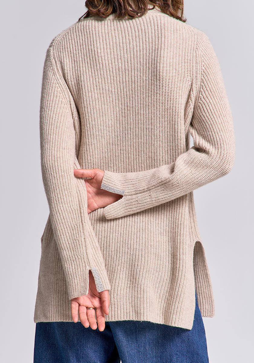 A person wearing a beige Princess Goes Hollywood Strickpullover aus Wolle-Kaschmir-Mix and blue jeans stands with their back to the camera, left arm bent behind them, both hands visible near the hem of the sweater.