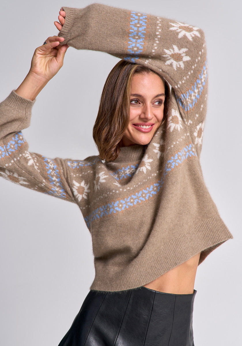 A smiling woman with shoulder-length brown hair raises her arms, revealing her waist in a beige Princess Goes Hollywood jacquard pullover with white and blue snowflake patterns and a black leather skirt, posing against a neutral gray background.