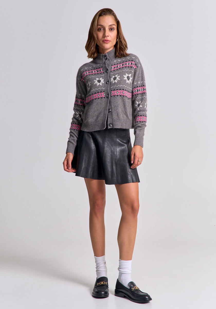 A woman faces forward in a gray Princess Goes Hollywood Fair Isle cardigan with pink and white Jacquard patterns, black leather skirt, white socks, and black loafers with gold chain detail. Her shoulder-length hair is styled in loose waves. Light gray background.