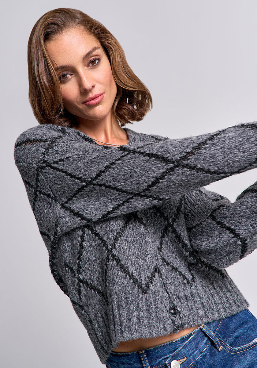 A woman with chin-length brown hair wears a textured gray Strickjacke by Princess Goes Hollywood featuring a black argyle pattern. She poses with arms outstretched, smiling slightly, in blue jeans against a plain light gray background.