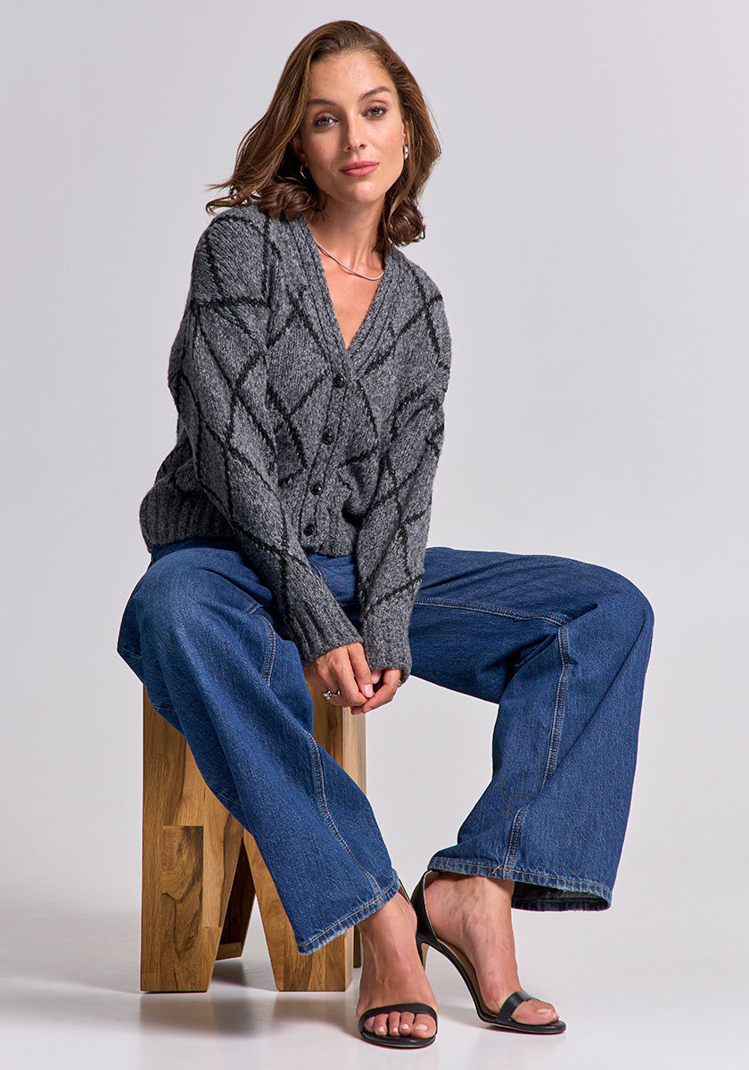 A woman with wavy brown hair sits calmly on a wooden stool against a plain background, wearing a Princess Goes Hollywood gray Argyle-Muster Strickjacke, blue wide-leg jeans, and black heeled sandals, her hands loosely clasped.