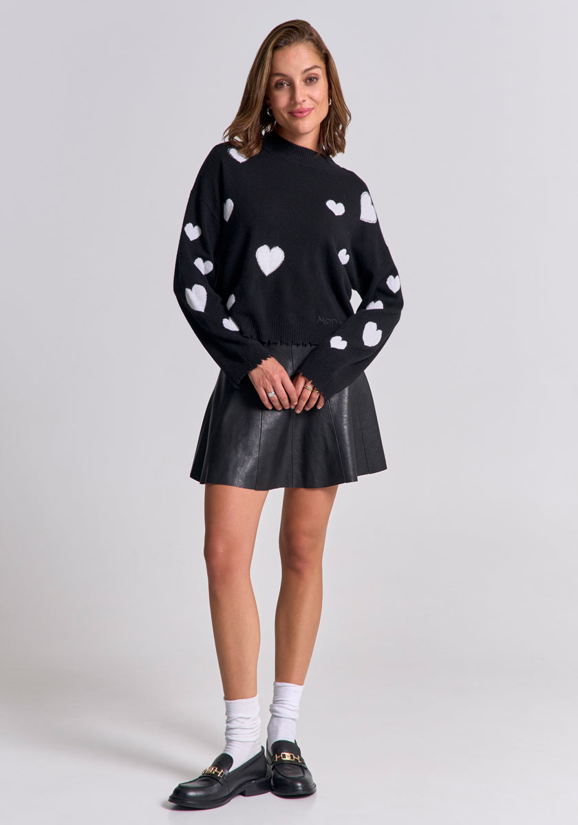A woman smiles in a Princess Goes Hollywood Feinstrickpullover mit Herz-Muster, a black faux leather skirt, white socks, and black loafers. She stands against a plain light gray background with her hands loosely clasped in front.