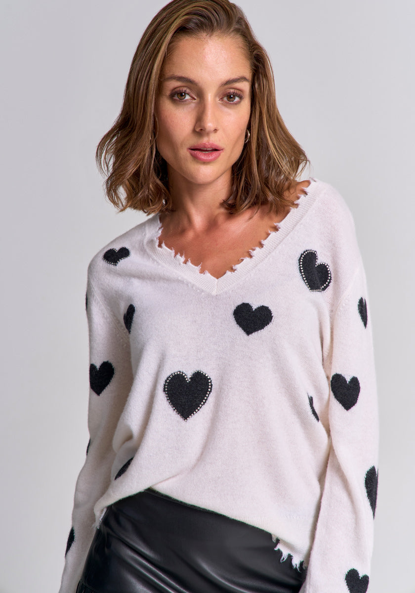 A woman with shoulder-length brown hair wears a white fuzzy heart-patterned fine-knit pullover by Princess Goes Hollywood with lightly frayed edges, paired with a black leather skirt. She stands against a light gray background and looks confidently at the camera.