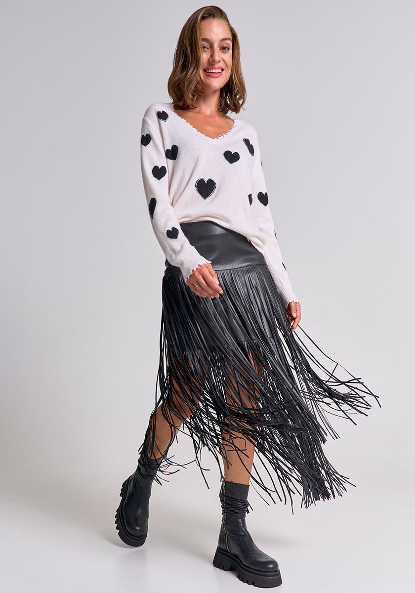 A woman with shoulder-length brown hair smiles in a Princess Goes Hollywood Feinstrickpullover mit Herz-Muster, black faux-leather fringe skirt, and black ankle boots. The moving fringes add a dynamic touch against the plain background.