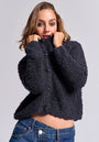 A person with light brown hair wears a textured black Princess Goes Hollywood Strickjacke aus Wolle-Alpaka-Mix and blue jeans, holding the collar up over their mouth while gazing neutrally at the camera against a plain light gray background.