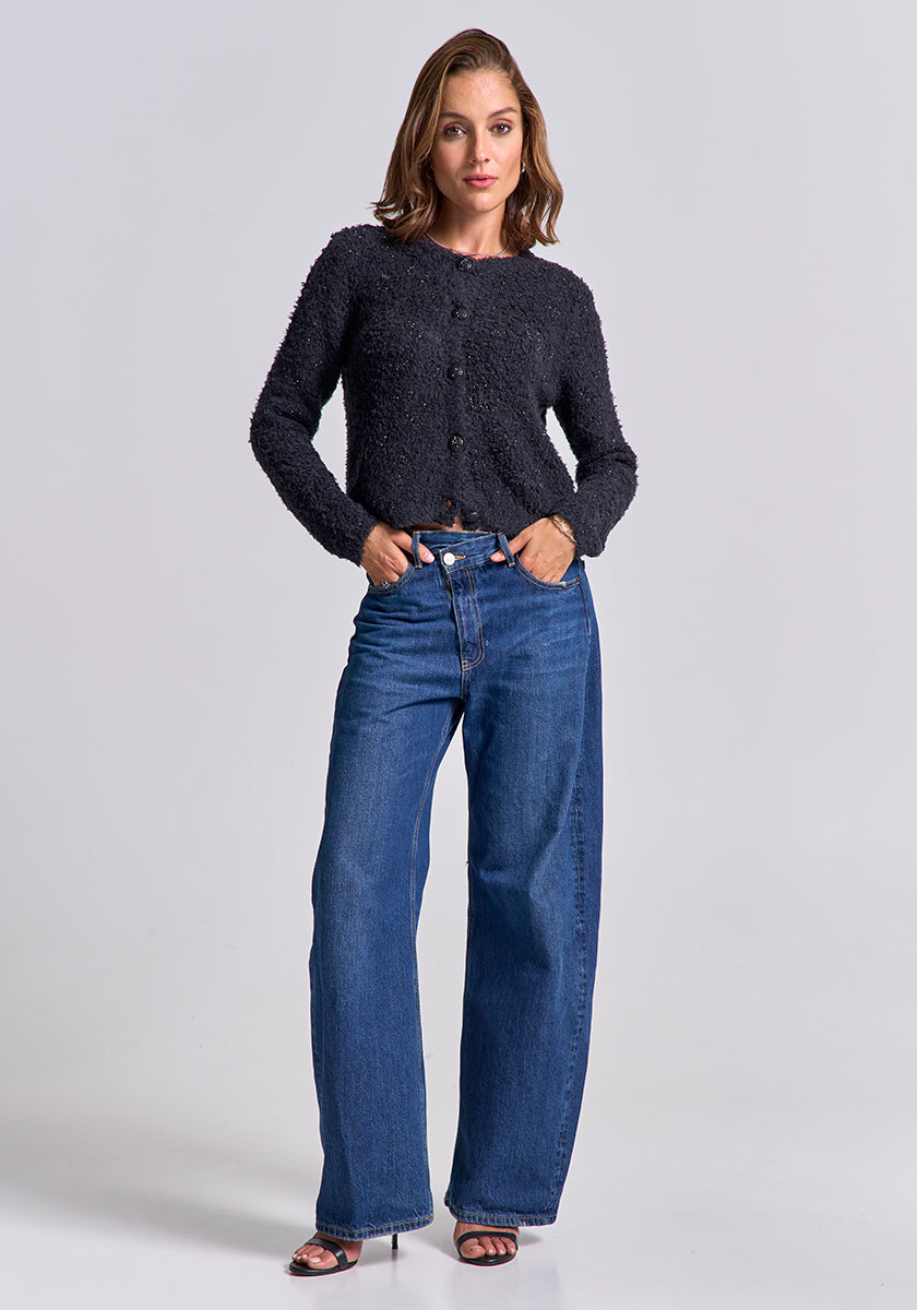A woman poses against a plain light gray background, wearing a textured zwarte Strickjacke aus Wolle-Alpaka-Mix by Princess Goes Hollywood with high-waisted, wide-leg blue jeans, black heeled sandals, and loose brown hair. One hand rests in her pocket.