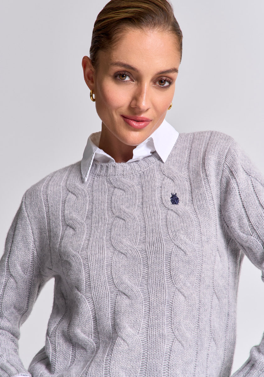 A woman with light makeup and slicked-back hair wears a light gray Princess Goes Hollywood Zopfstrick Wolle-Kaschmir-Mix Pullover with a round neckline over a white collared shirt, accessorized with gold hoop earrings, against a plain light gray background.