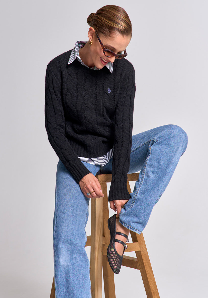 A woman in a Princess Goes Hollywood schwarzen Zopfstrick Wolle-Kaschmir-Mix Pullover mit Rundhalsausschnitt over a striped shirt and blue jeans sits on a wooden stool, smiling as she adjusts her black mesh flat shoe, glasses and gold hoop earrings framing her short hair.