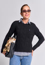 A smiling woman wears Princess Goes Hollywood cat-eye sunglasses, gold hoop earrings, a black Zopfstrick Wolle-Kaschmir-Mix Pullover, a striped collared shirt, blue jeans, and holds a black handbag with a patterned scarf against a plain background.