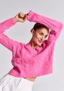 A woman in a bright pink Princess Goes Hollywood Zopfstrick Pullover aus Wolle-Kaschmir and white pants poses with arms raised and crossed above her head, slightly smiling. She wears gold hoop earrings, with short brown hair pulled back, on a light gray background.