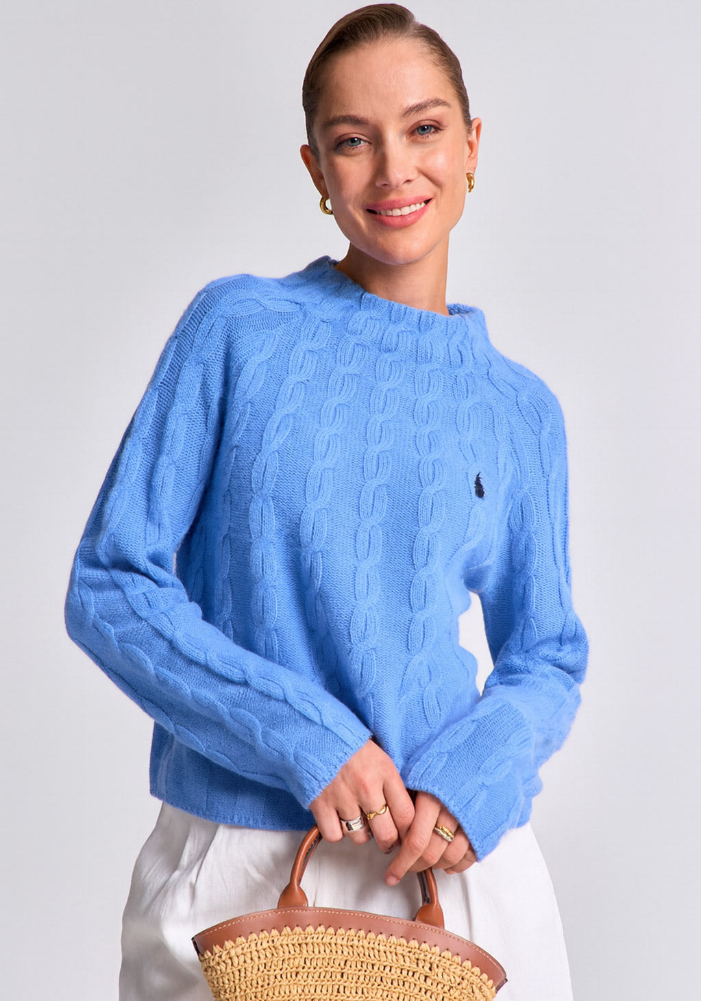 A woman with light skin and slicked-back hair smiles softly, wearing a light blue Princess Goes Hollywood Zopfstrick Pullover Wolle-Kaschmir-Mix with Stehkragen and white pants, holding a woven straw bag with brown leather handle against a plain light gray background.