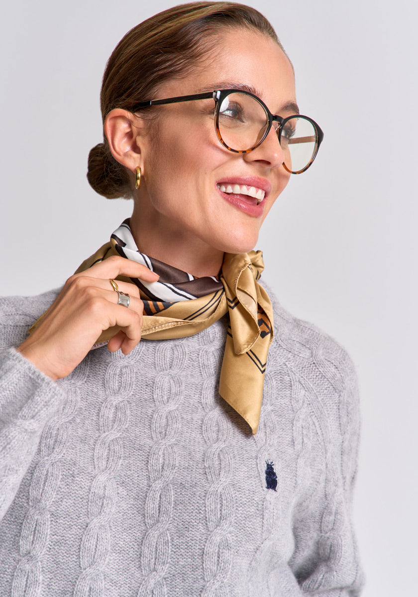A smiling woman with slicked-back hair wears large round glasses, a gold hoop earring, a pale gold patterned scarf, and the light gray Zopfstrick pullover with logo and stand-up collar by Princess Goes Hollywood.