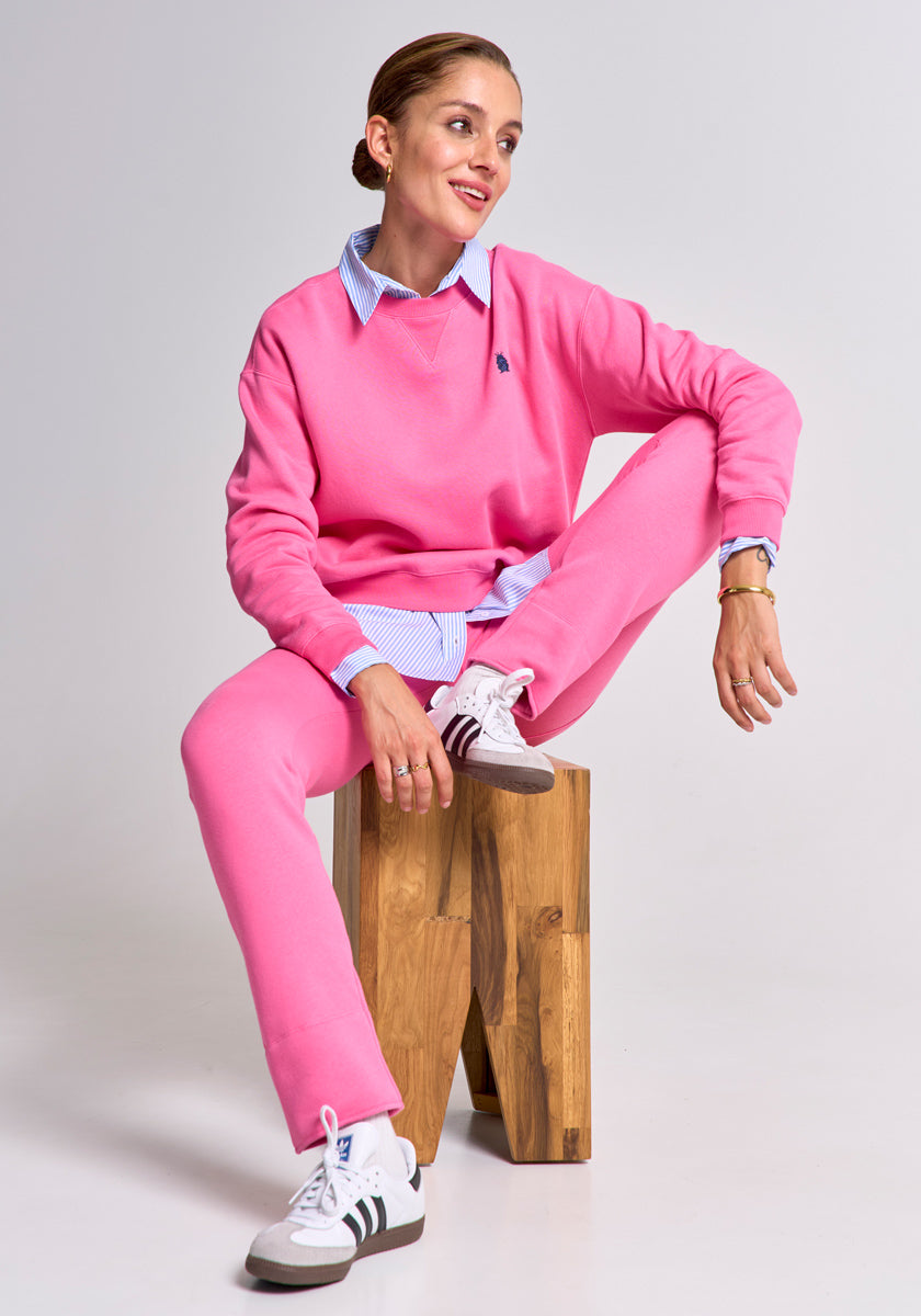 A woman sits on a wooden block against a plain background, wearing a bright pink Regular Fit sweatshirt and Jogginghose by Princess Goes Hollywood over a blue striped collared shirt, paired with white sneakers. Her hair is tied back as she smiles gently to the side.