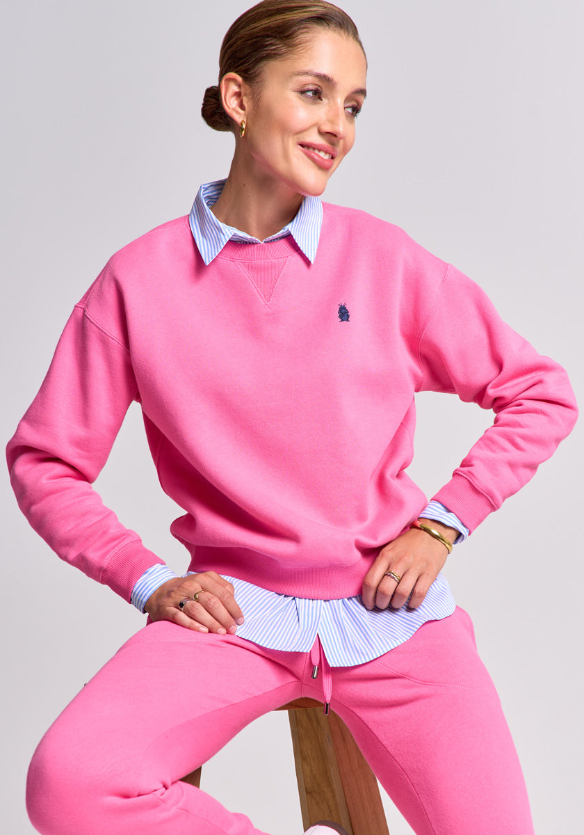 A woman sits on a wooden stool, smiling with her hair pulled back, wearing a bright pink Princess Goes Hollywood sweatpullover and matching pants over a blue and white striped button-down shirt. Her hands rest on her knees against a light background.