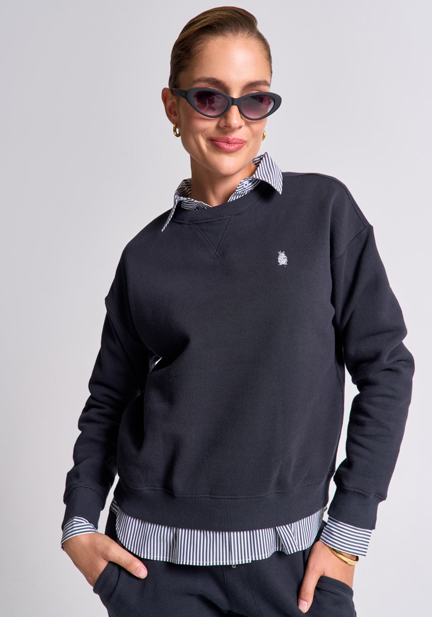 A person with slicked-back hair wears cat-eye sunglasses, hoop earrings, and a black Princess Goes Hollywood Sweatpullover aus Baumwolle over a striped button-up shirt. They pose confidently with one hand in their pocket against a light gray background.