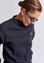 A woman with slicked-back hair in a low bun wears black rectangular sunglasses, gold hoop earrings, and a dark cotton Sweatpullover with a small embroidered logo by Princess Goes Hollywood. She looks down neutrally against a plain light background.