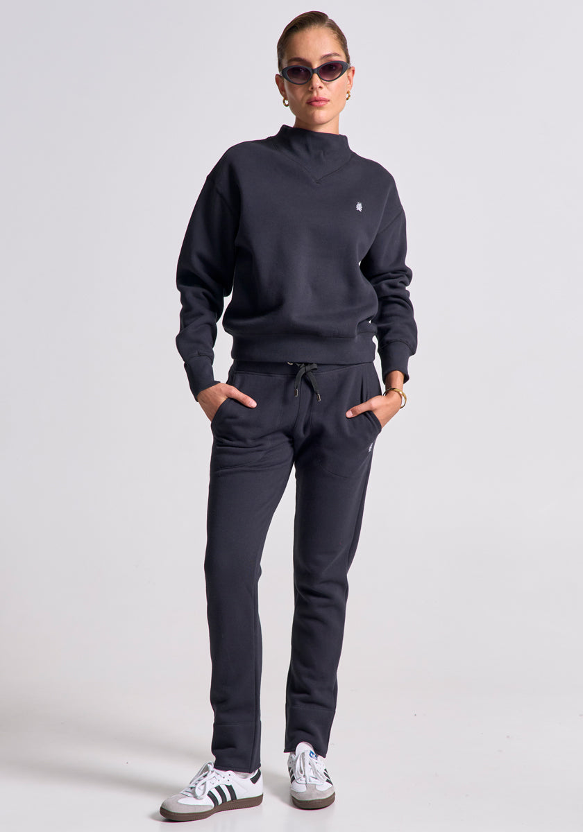 A woman faces the camera with hands in pockets, wearing a black Princess Goes Hollywood Regular Fit sweatshirt and cotton joggers, white Adidas sneakers, dark sunglasses, and a watch. Her hair is tied back against a plain light background.