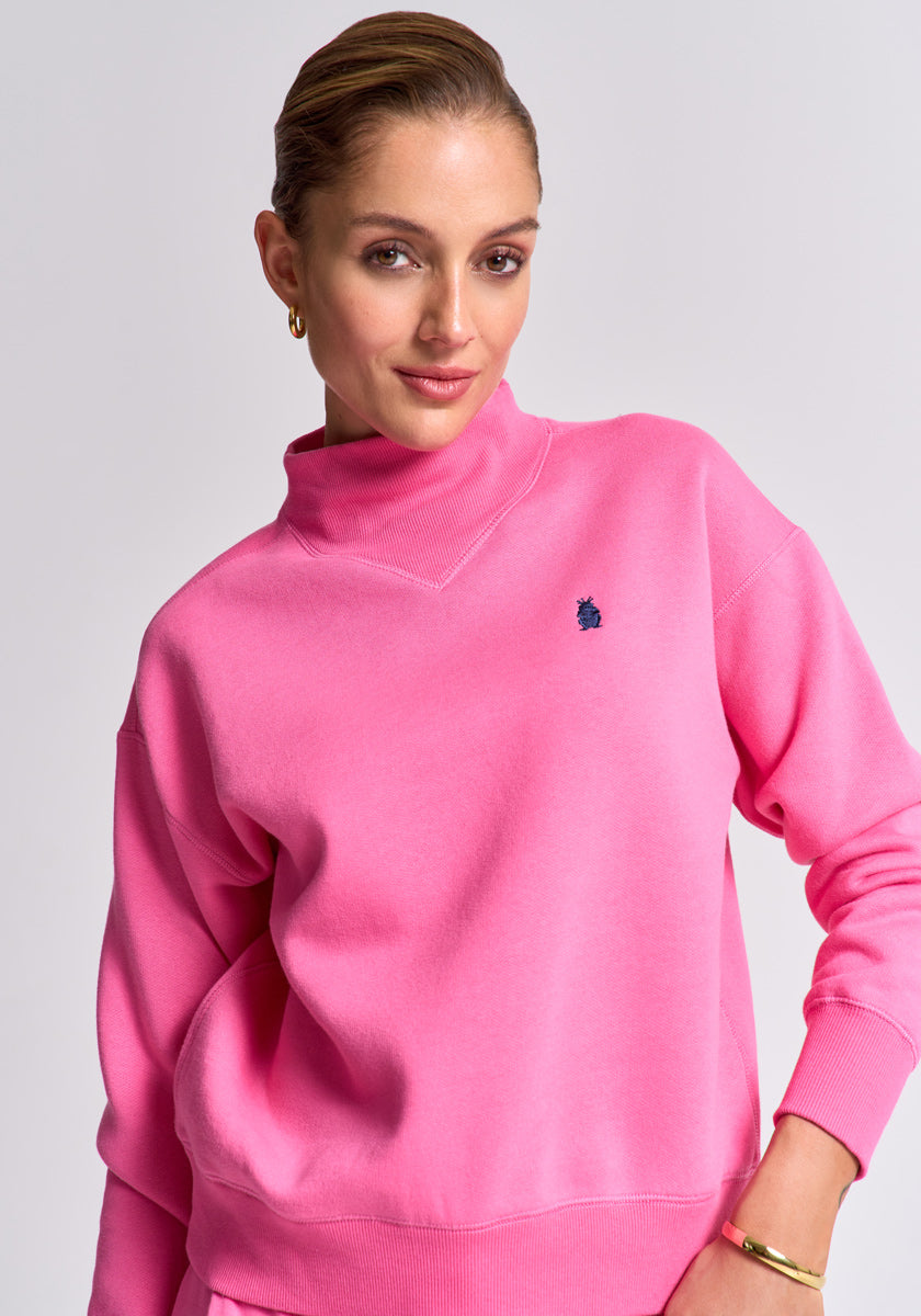 A woman with light skin and brown hair tied back wears a bright pink Princess Goes Hollywood Baumwolle Sweatpullover featuring a small navy blue embroidered logo on the chest, paired with gold hoop earrings and bracelet, standing against a light gray background.
