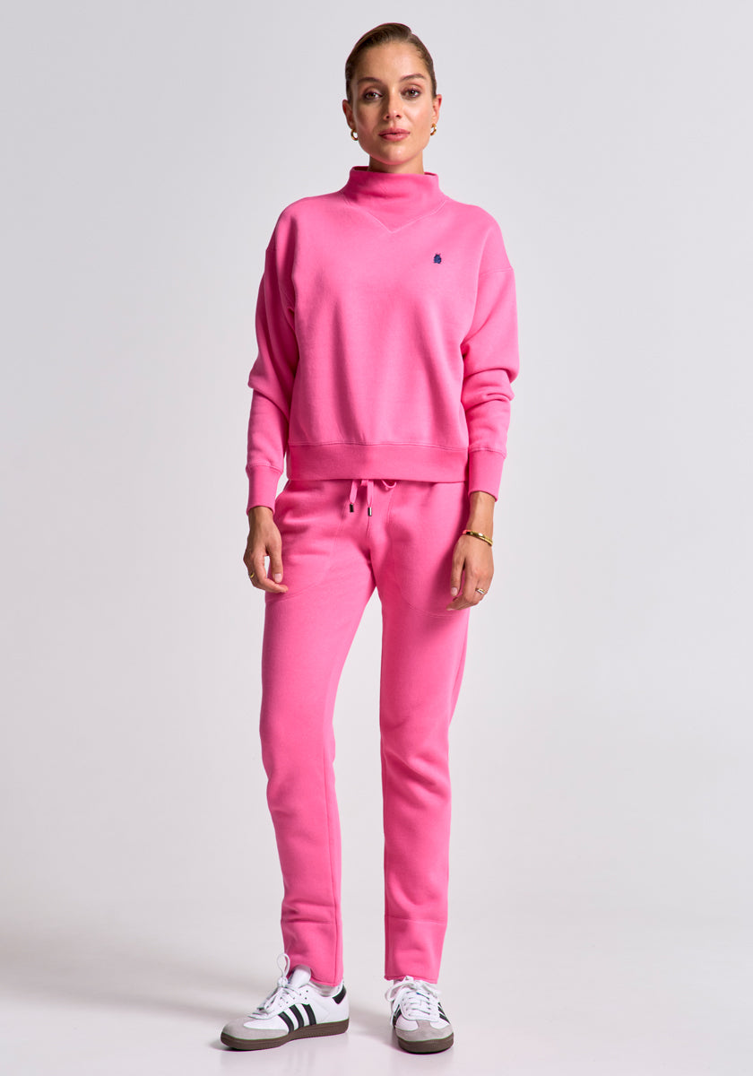 A woman with light skin and hair tied back wears a bright pink Princess Goes Hollywood Sweatpullover and sweatpants set in soft Baumwolle-Fleece, paired with white sneakers featuring black stripes, against a plain light gray background.
