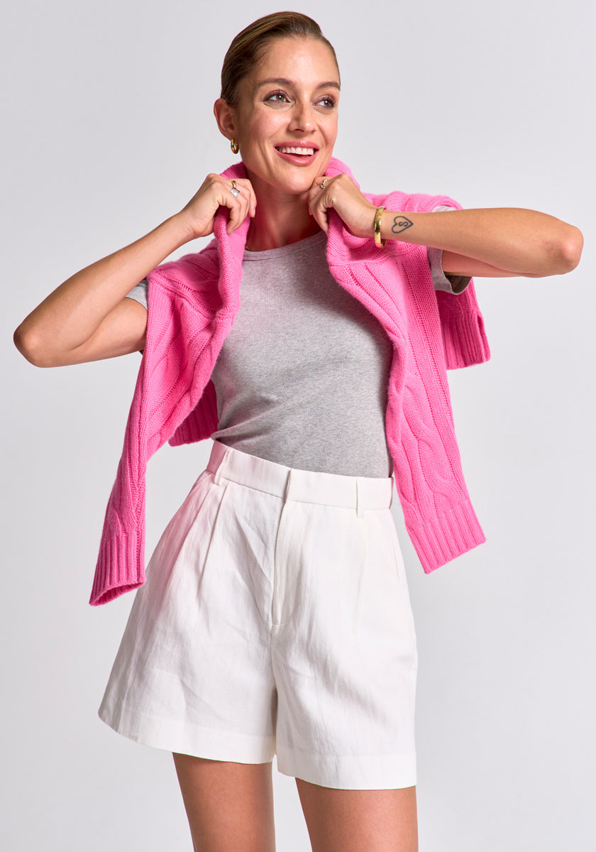 A smiling woman poses against a plain background in a Princess Goes Hollywood graues Baumwoll-T-Shirt mit Rundhalsausschnitt, das in ihre hochgeschnittenen weißen Shorts gesteckt ist, mit pinkem Pullover über den Schultern und eleganten Accessoires.