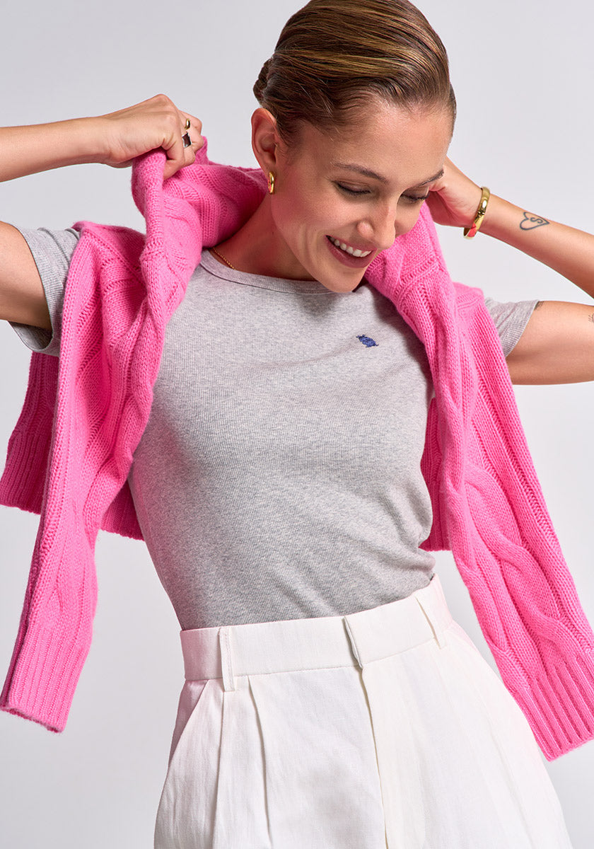 A smiling person with slicked-back hair wears a grey Princess Goes Hollywood Baumwoll-T-Shirt mit Rundhalsausschnitt, white high-waisted pants, and a bright pink sweater over their shoulders, accessorized with gold hoops, a bracelet, and a small heart tattoo.