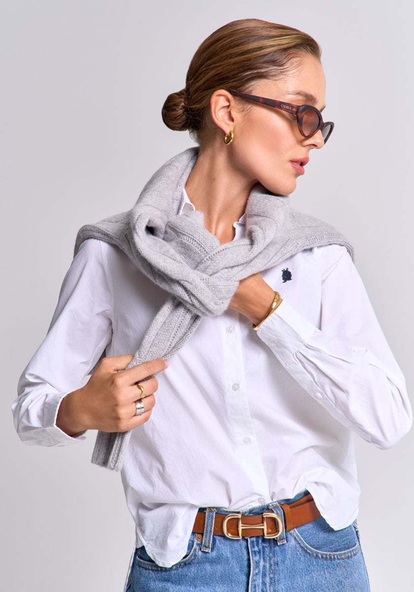 A woman with short, slicked-back hair wears sunglasses, gold hoops, blue jeans, a brown belt, rings, a gold bracelet, and has a Princess Goes Hollywood Pullover Wolle-Kaschmir mit V-Ausschnitt draped over her shoulders as she looks left.